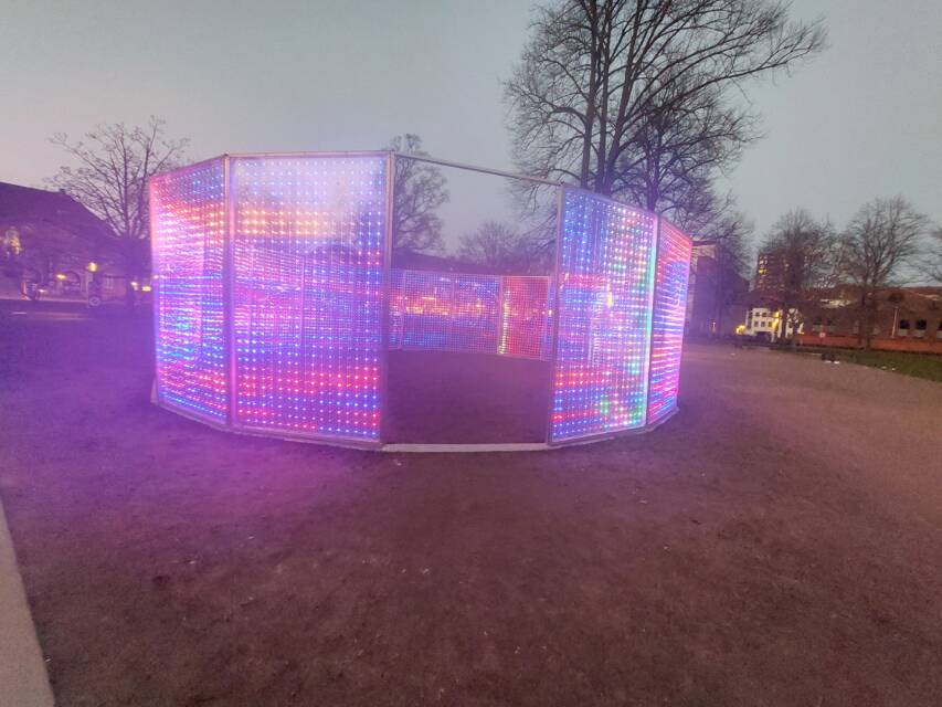 Installation artistique sur une place publique de forme octogonale couverte de LEDs multicolores photographiée au crépuscule